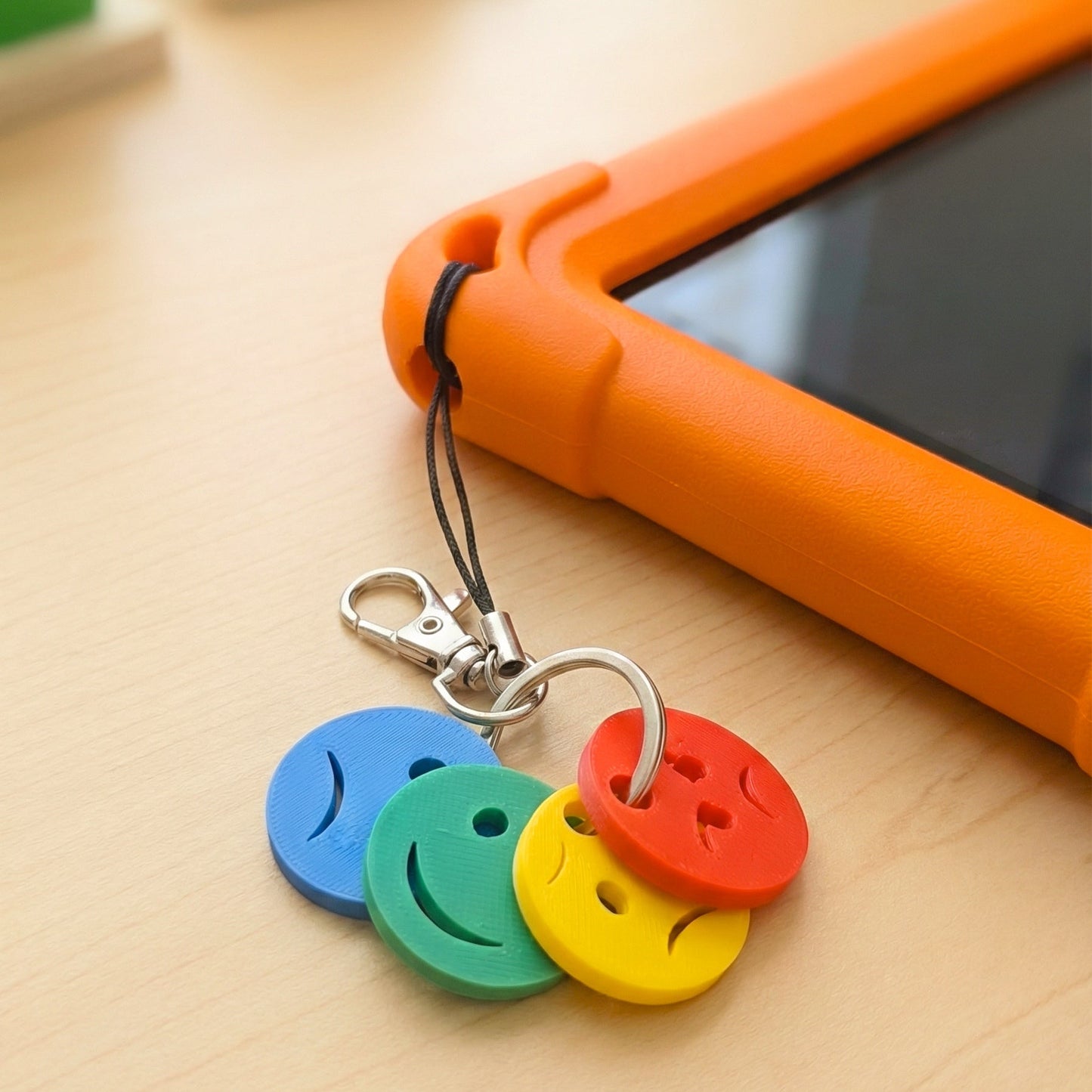 A detailed close-up of the Emotions Device Charm for phones and tablets, featuring four textured, colorful discs with various facial expressions attached via a black loop to the corner of a rugged orange child-friendly tablet case