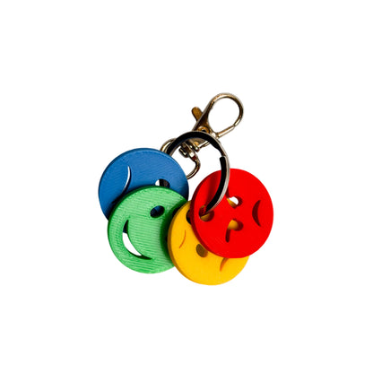 E-commerce infographic featuring a close-up of the Mini Emotions Keychain with four 3D-printed faces in blue, green, yellow, and red, attached to a metal clip. Below the product, three icons indicate benefits: 'TAKE IT ANYWHERE', 'DURABLE & FUN DESIGN', and 'INCLUDES CLIP'.