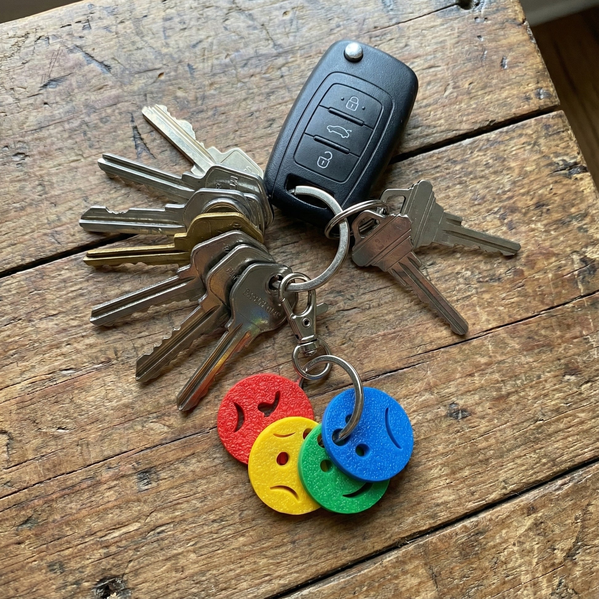 Top-down product photograph showing the mini emotions keychain with four colored discs (red, yellow, green, blue) attached to a large bunch of standard metal keys and a car fob, resting flat on a rustic wooden table. This overhead view demonstrates the small scale of the discs compared to everyday keys.